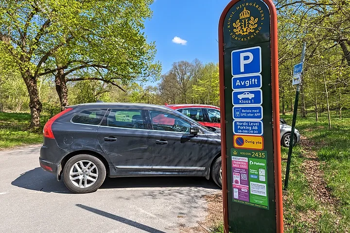 Parkeringsskylt med Kungliga Djurgårdens förvaltnings emblem, P-skylt och parkeringsbolagets namn, bredvid parkerade bilar
