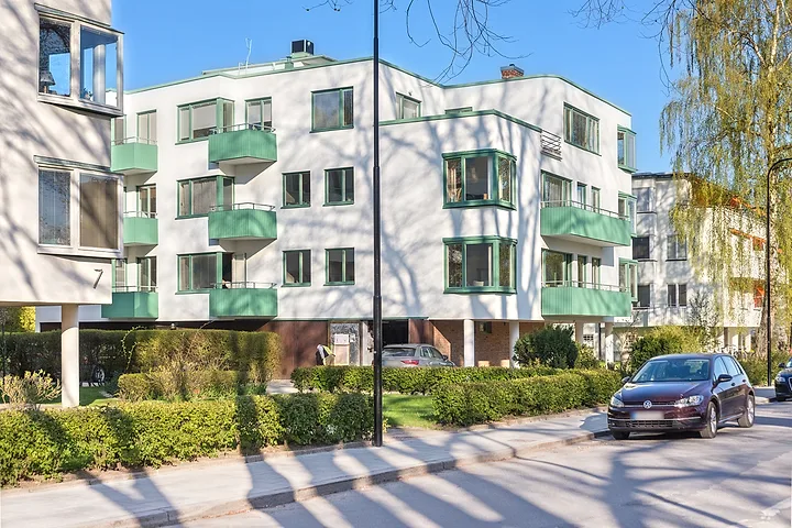 Vitt flerbostadshus med gröna balkonger och en bil parkerad längs gatan