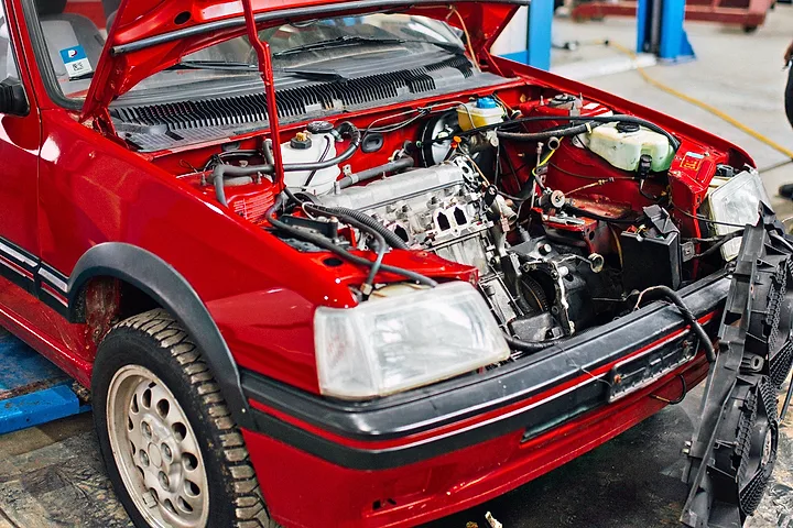 Röd Peugeot 205 med öppen motorhuv och delvis demonterad front i en verkstad